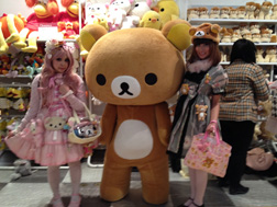 Rilakkuma during a launch event at FAO Schwarz in New York City, flanked by AQI representatives Julie (left) and Stephanie (right), who are dressed in Lolita fashion.
